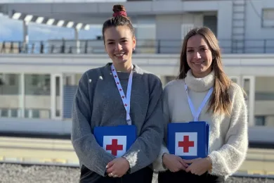 Zwei junge Frauen stehen mit Unterlagen vom Roten Kreuz vor einem Gebäude.
