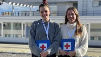 Zwei junge Frauen stehen mit Unterlagen vom Roten Kreuz vor einem Gebäude.