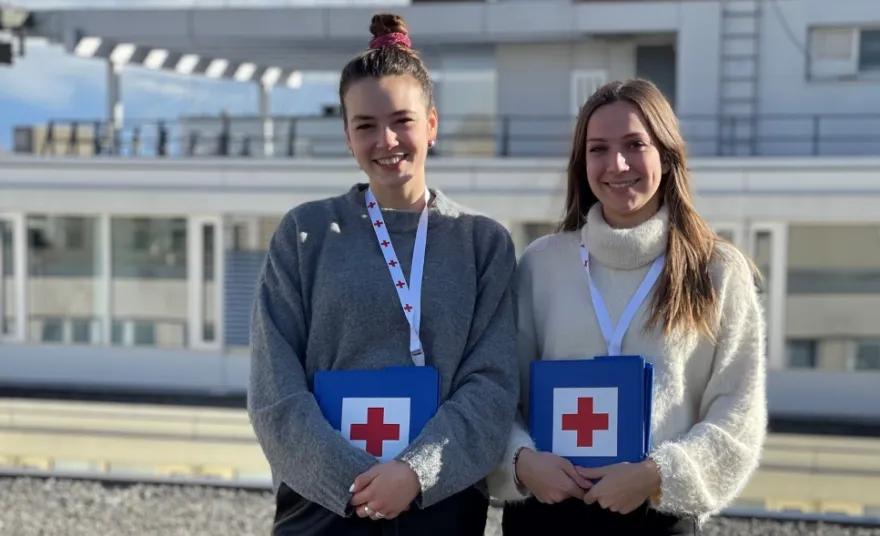 Zwei junge Frauen stehen mit Unterlagen vom Roten Kreuz vor einem Gebäude.